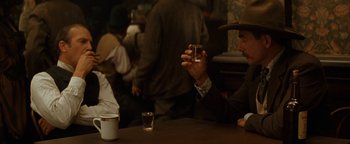 Movie still from “Wyatt Earp” (1994), directed by Lawrence Kasdan – A man sitting at a table holding a glass of beer; Medium shot, Over the shoulder angle