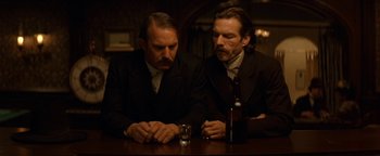 Movie still from “Wyatt Earp” (1994), directed by Lawrence Kasdan – A couple of men sitting at a table with a bottle of alcohol; Medium shot, Low angle
