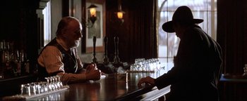 Movie still from “Wyatt Earp” (1994), directed by Lawrence Kasdan – Two men sitting at a table in front of each other at a bar; Medium shot, Over the shoulder angle