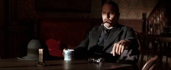 Movie still from “Wyatt Earp” (1994), directed by Lawrence Kasdan – A man sitting at a table smoking a cigarette and holding a cup; Medium shot, Low angle