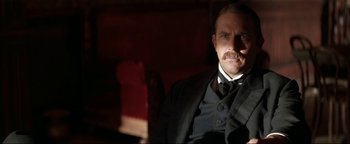Movie still from “Wyatt Earp” (1994), directed by Lawrence Kasdan – A man in a suit and tie sitting in a chair; Close Up shot, Low angle