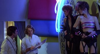 Movie still from “Xanadu” (1980), directed by Robert Greenwald – A man and a woman are standing in front of a surfboard; Medium shot, Low angle