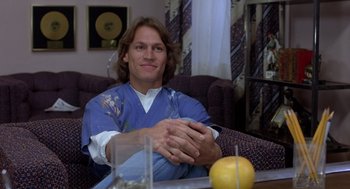 Movie still from “Xanadu” (1980), directed by Robert Greenwald – A man sitting at a coffee table with his hands clasped together; Medium shot, Low angle