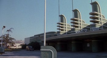 Movie still from “Xanadu” (1980), directed by Robert Greenwald – A car is parked on the side of the road near a building; Extreme Wide shot, Low angle