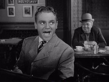 Movie still from “Yankee Doodle Dandy” (1942), directed by Michael Curtiz – A man in a suit and hat sitting at a table with an older man; Medium shot, Over the shoulder angle