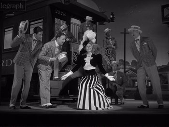 Movie still from “Yankee Doodle Dandy” (1942), directed by Michael Curtiz – A group of people standing around a woman in a striped dress; Wide shot, Low angle