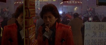 Movie still from “Year of the Dragon” (1985), directed by Michael Cimino – A man smoking a cigarette in a crowded area; Medium shot, Over the shoulder angle