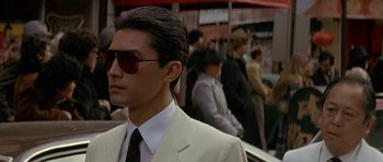Movie still from “Year of the Dragon” (1985), directed by Michael Cimino – A man wearing a suit and sunglasses in front of a group of people; Close Up shot, Over the shoulder angle