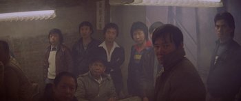 Movie still from “Year of the Dragon” (1985), directed by Michael Cimino – A group of people standing around in a room; Medium shot, High angle