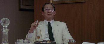 Movie still from “Year of the Dragon” (1985), directed by Michael Cimino – A man in a white suit sitting in front of a plate of food; Medium shot, Low angle