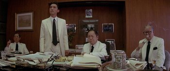 Movie still from “Year of the Dragon” (1985), directed by Michael Cimino – A man sitting at a desk in front of another man in a suit; Medium shot, Low angle