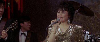 Movie still from “Year of the Dragon” (1985), directed by Michael Cimino – A woman is singing into a microphone in front of a crowd; Close Up shot, Low angle