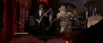 Movie still from “Year of the Dragon” (1985), directed by Michael Cimino – A group of people that are singing in a room; Medium shot, High angle