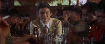 Movie still from “Year of the Dragon” (1985), directed by Michael Cimino – A man sitting at a table in front of a bunch of bottles; Medium shot, Over the shoulder angle