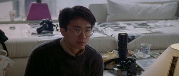 Movie still from “Year of the Dragon” (1985), directed by Michael Cimino – A man with glasses and a sweater sitting in a room with a camera; Close Up shot, High angle