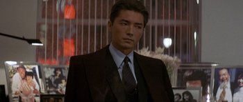 Movie still from “Year of the Dragon” (1985), directed by Michael Cimino – A man wearing a suit and tie standing in front of flowers; Close Up shot, Over the shoulder angle