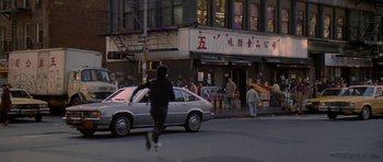 Movie still from “Year of the Dragon” (1985), directed by Michael Cimino – A white car driving down a street next to a building; Wide shot, High angle