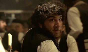 Movie still from “Yentl” (1983), directed by Barbra Streisand – A man with a beard and curly hair in front of a group of people; Close Up shot, Over the shoulder angle