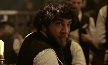 Movie still from “Yentl” (1983), directed by Barbra Streisand – A man with a beard and curly hair in front of a group of people; Close Up shot, Over the shoulder angle