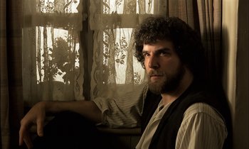 A man sitting in front of a window with lace curtains