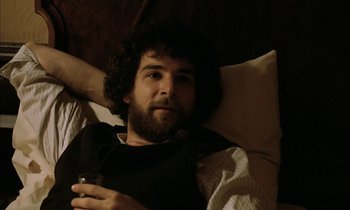 Movie still from “Yentl” (1983), directed by Barbra Streisand – A bearded man laying in a bed holding a drink; Close Up shot, High angle