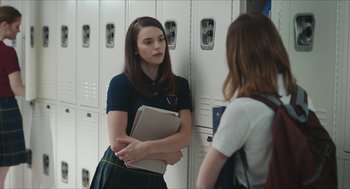 Movie still from “Yes, God, Yes” (2019), directed by Karen Maine – A girl in a school uniform is holding a book and talking to another girl in front of a lockers; Medium shot, Over the shoulder angle
