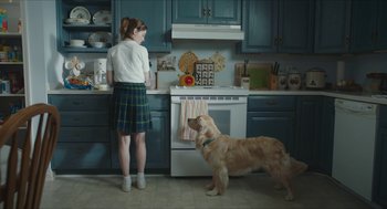 Movie still from “Yes, God, Yes” (2019), directed by Karen Maine – A woman and a dog standing in a kitchen next to a stove; Wide shot, Over the shoulder angle