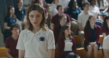 Movie still from “Yes, God, Yes” (2019), directed by Karen Maine – A girl in a white shirt is standing in front of a group of people; Medium shot, Over the shoulder angle