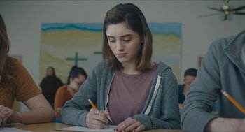 Movie still from “Yes, God, Yes” (2019), directed by Karen Maine – A girl is writing on a piece of paper; Medium shot, Over the shoulder angle