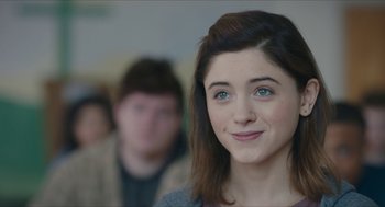 Movie still from “Yes, God, Yes” (2019), directed by Karen Maine – A young woman smiles for the camera while a young man looks on; Close Up shot, Over the shoulder angle