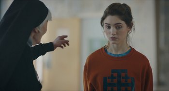 Movie still from “Yes, God, Yes” (2019), directed by Karen Maine – A woman standing next to another woman in an orange shirt; Close Up shot, Over the shoulder angle