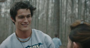 Movie still from “Yes, God, Yes” (2019), directed by Karen Maine – A young man smiling in the woods while wearing a necklace; Close Up shot, Over the shoulder angle