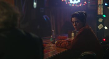 Movie still from “Yes, God, Yes” (2019), directed by Karen Maine – A woman sitting at a table with a bottle of beer; Medium shot, Over the shoulder angle