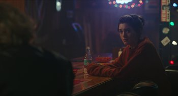 Movie still from “Yes, God, Yes” (2019), directed by Karen Maine – A woman sitting at a table with a bottle of beer; Medium shot, Over the shoulder angle