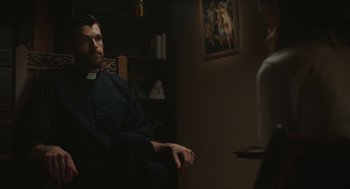 Movie still from “Yes, God, Yes” (2019), directed by Karen Maine – A man sitting in a chair in front of a woman; Medium shot, Over the shoulder angle