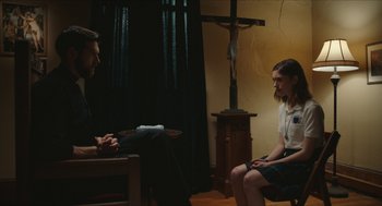 Movie still from “Yes, God, Yes” (2019), directed by Karen Maine – A man and a woman sitting on a chair in front of a crucifix; Medium shot, Over the shoulder angle