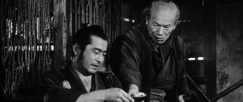 Movie still from “Yojimbo” (1961), directed by Akira Kurosawa – Two men sitting in front of a bamboo wall; Medium shot, Low angle