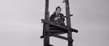 Movie still from “Yojimbo” (1961), directed by Akira Kurosawa – A black and white photo of a man sitting on a wooden structure; Wide shot, Low angle