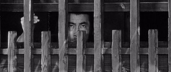 Movie still from “Yojimbo” (1961), directed by Akira Kurosawa – A black and white photo of a man looking through a fence; Close Up shot, Overhead angle