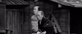 Movie still from “Yojimbo” (1961), directed by Akira Kurosawa – A man riding a bike down a street with another man standing behind him; Medium shot, Low angle