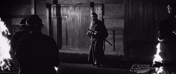 Movie still from “Yojimbo” (1961), directed by Akira Kurosawa – A man holding a sword in front of a wooden wall; Wide shot, Over the shoulder angle