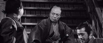 Movie still from “Yojimbo” (1961), directed by Akira Kurosawa – An older man in a kimono sitting in front of a wooden wall; Medium shot, Low angle