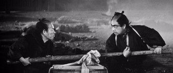 Movie still from “Yojimbo” (1961), directed by Akira Kurosawa – Two men in traditional garb sit in front of a wooden box; Medium shot, Over the shoulder angle