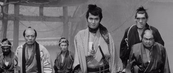 Movie still from “Yojimbo” (1961), directed by Akira Kurosawa – A man holding a drink in his hand while standing next to another man; Medium shot, Over the shoulder angle