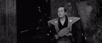 Movie still from “Yojimbo” (1961), directed by Akira Kurosawa – A man holding a racket in front of a wooden wall; Medium shot, Low angle