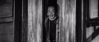 Movie still from “Yojimbo” (1961), directed by Akira Kurosawa – A black - and - white photo of a man smiling in a doorway; Medium shot, Low angle