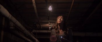 Movie still from “You're Next” (2011), directed by Adam Wingard – A woman standing under a light in a dark room; Medium shot, Low angle