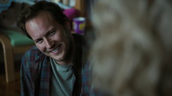 Movie still from “Young Adult” (2011), directed by Jason Reitman – A man smiling for the camera while sitting in a room; Close Up shot, Over the shoulder angle