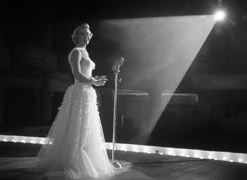Movie still from “Young Man with a Horn” (1950), directed by Michael Curtiz – A woman in a white dress standing in front of a microphone; Wide shot, Low angle