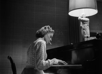Movie still from “Young Man with a Horn” (1950), directed by Michael Curtiz – A woman sitting at a piano with a cigarette in her mouth; Medium shot, Low angle